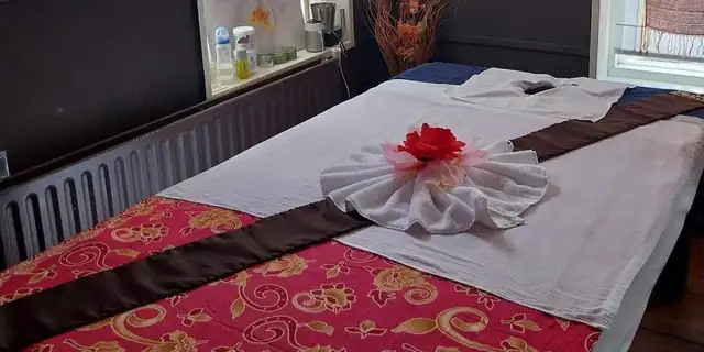 Behandelruimte van PheunThai Massage in Makkinga met een massagetafel onder een rood kleed met gouden bloemen, een handdoek in de vorm van een bloem en daglicht door het raam