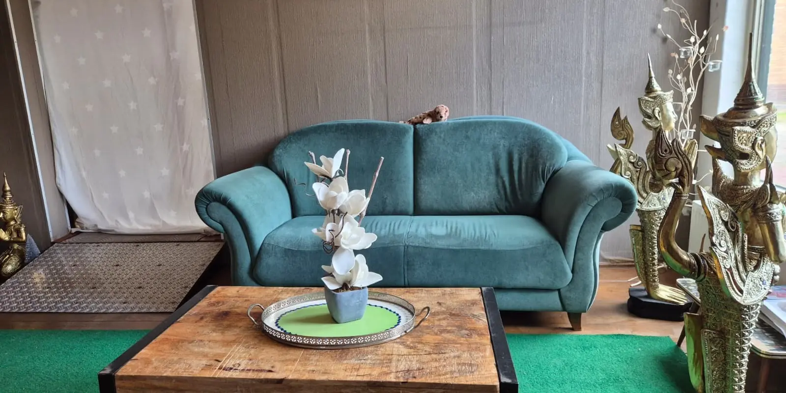 Zithoek in de salon van PheunThai Massage in Makkinga met een groene bank, een houten salontafel en Thaise beelden